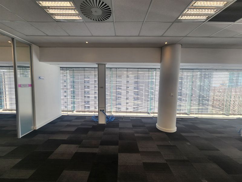 A bright, open-plan office space featuring carpet tiles, suspended ceilings with integrated lighting, and large floor-to-ceiling windows with venetian blinds.