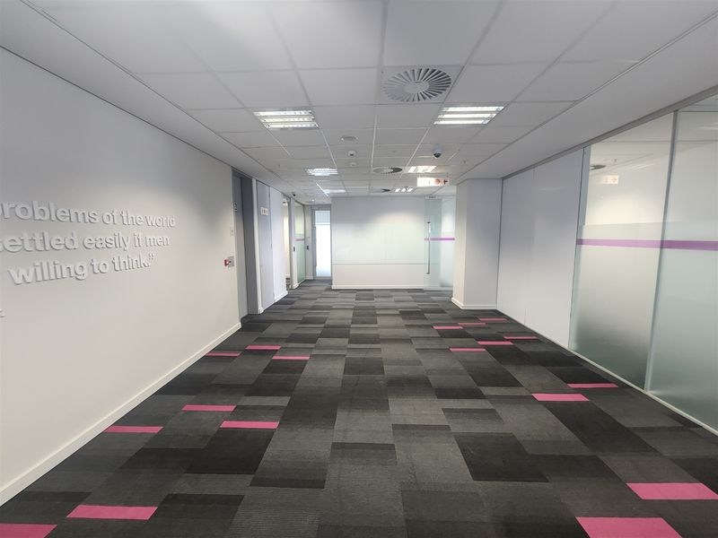 A modern, well-maintained office hallway featuring carpet tiles, suspended ceilings with recessed lighting, and glass partition walls.