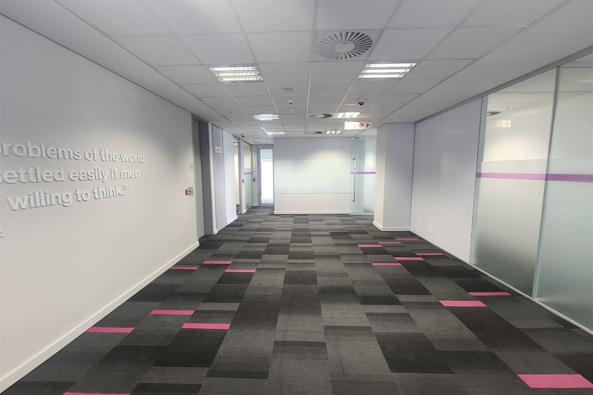 A modern, well-maintained office hallway featuring carpet tiles, suspended ceilings with recessed lighting, and glass partition walls.