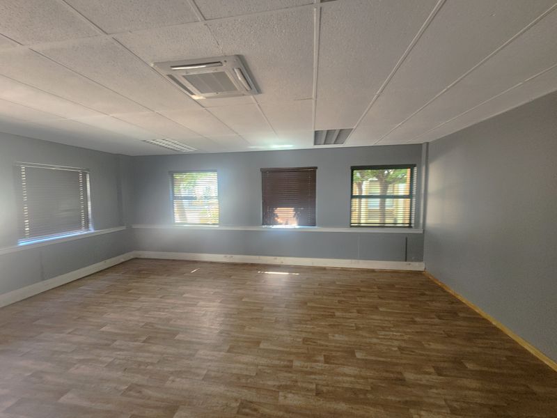 A spacious, empty office area featuring wood-look vinyl flooring, a suspended ceiling with recessed lighting, and multiple windows with fitted blinds.