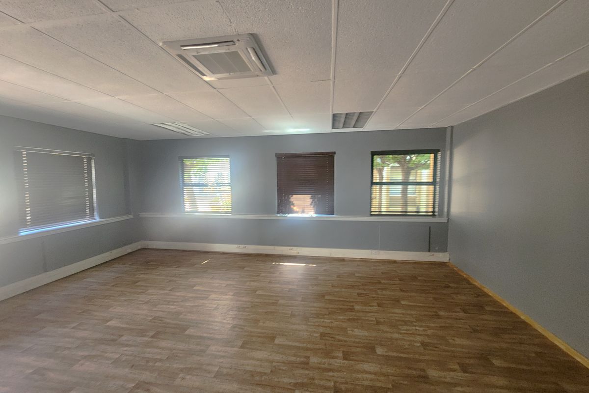 A spacious, empty office area featuring wood-look vinyl flooring, a suspended ceiling with recessed lighting, and multiple windows with fitted blinds.