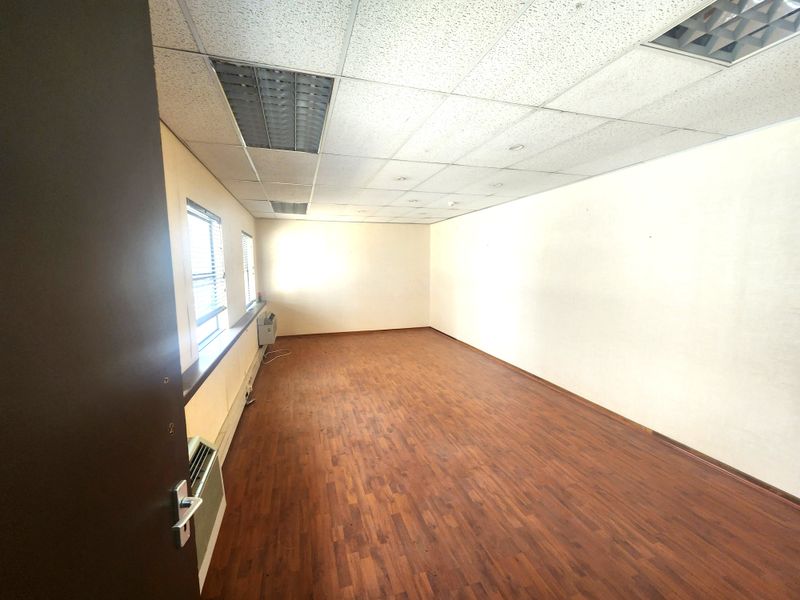 An empty office space featuring laminate flooring, a suspended ceiling with recessed lighting, and a built-in window air conditioning unit.