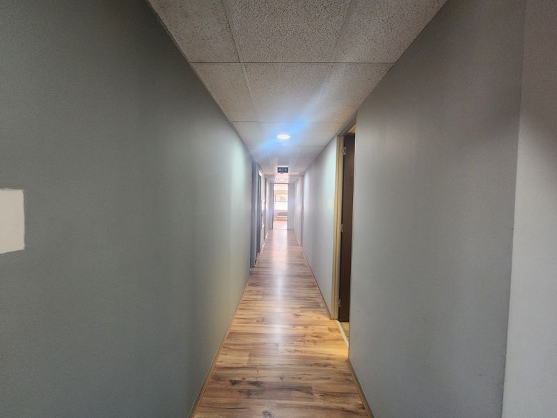 A clean, functional office hallway featuring wood-look laminate flooring, a suspended ceiling with recessed lighting, and neutral grey walls.