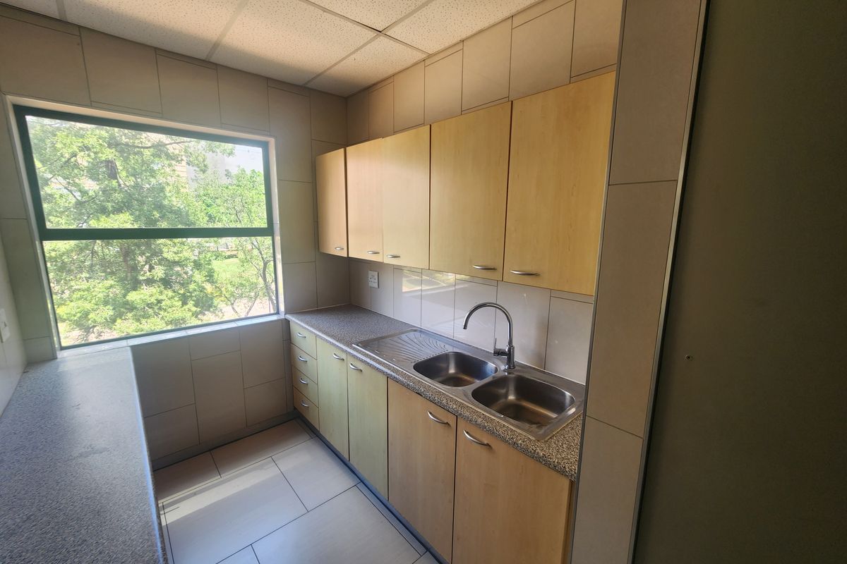 A functional office kitchenette featuring laminate cabinetry, a stainless steel double sink, tiled backsplash, and large window providing natural light.