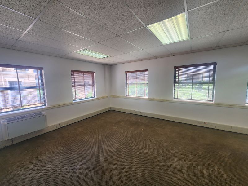 An empty office space featuring a suspended ceiling, recessed lighting, wall-mounted air conditioning, and carpet flooring with multiple windows providing natural light.