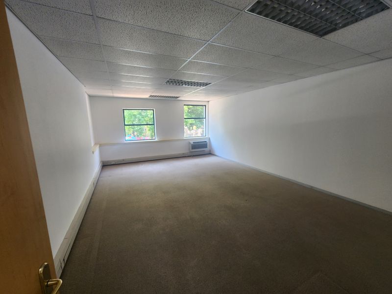 An empty, open-plan office space featuring a suspended ceiling with recessed lighting, fitted carpeting, and a wall-mounted air conditioning unit.