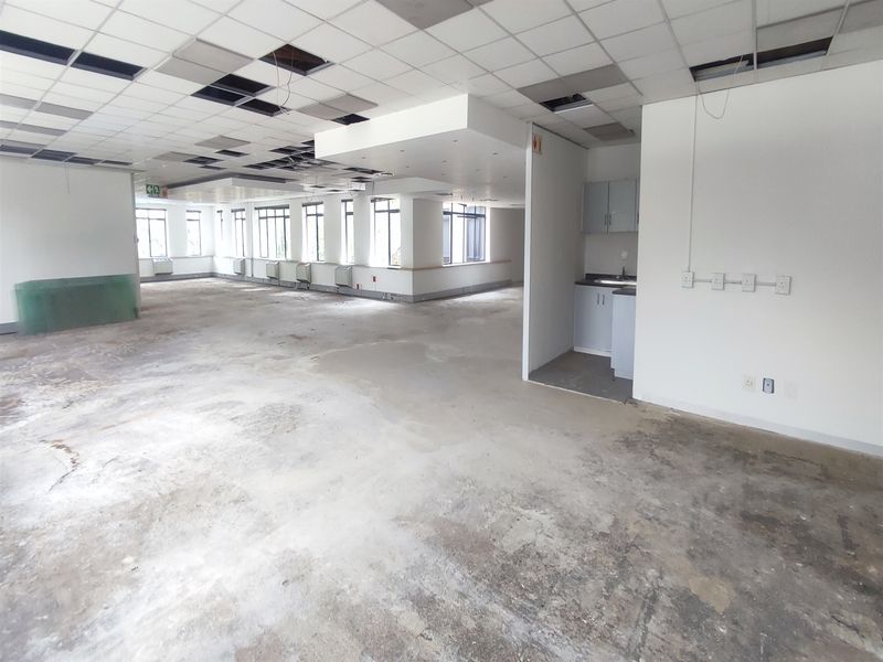 Open plan office space featuring large windows, a small kitchenette, and a suspended ceiling requiring maintenance and flooring repairs.