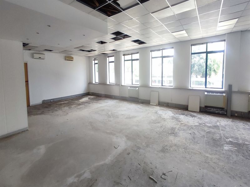 A vacant office space featuring large windows, split-system air conditioning, and a suspended ceiling grid with several missing tiles and unfinished flooring.