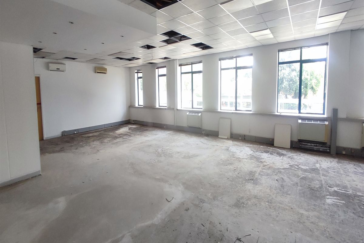 A vacant office space featuring large windows, split-system air conditioning, and a suspended ceiling grid with several missing tiles and unfinished flooring.