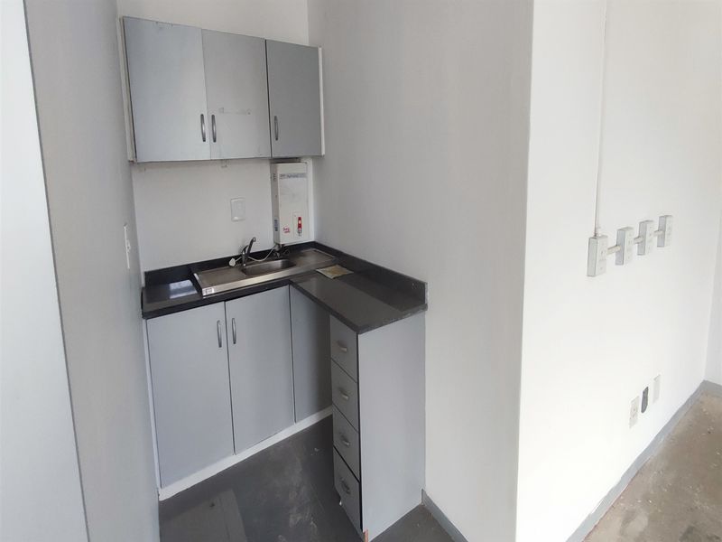 A compact office kitchenette featuring grey built-in cabinetry, a stainless steel sink, and a wall-mounted water heater in a functional, empty workspace.