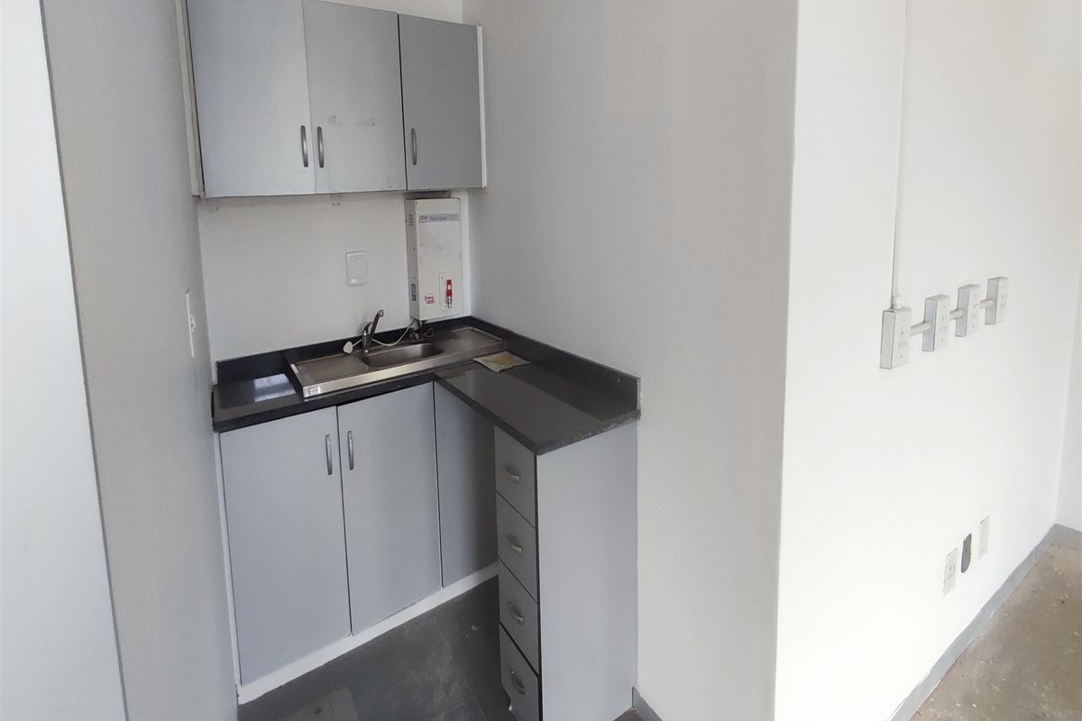 A compact office kitchenette featuring grey built-in cabinetry, a stainless steel sink, and a wall-mounted water heater in a functional, empty workspace.