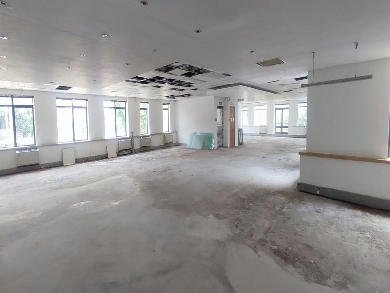 A large, open-plan office space featuring extensive natural light, suspended ceiling grid, and elevator access, currently requiring significant refurbishment and flooring repairs.