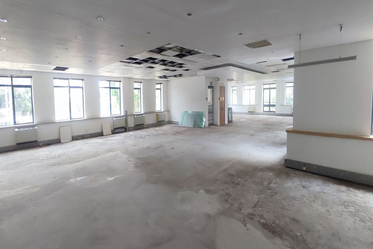 A large, open-plan office space featuring extensive natural light, suspended ceiling grid, and elevator access, currently requiring significant refurbishment and flooring repairs.