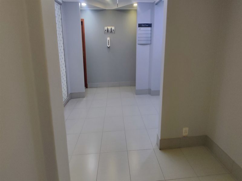 A clean, well-lit office hallway featuring neutral ceramic floor tiles, recessed ceiling lighting, and clear directional signage for the first floor.
