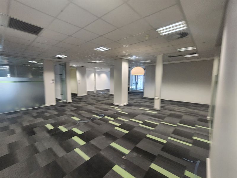 A spacious, open-plan office area featuring suspended ceilings, recessed lighting, and modern patterned carpet tiles with glass partition walls.
