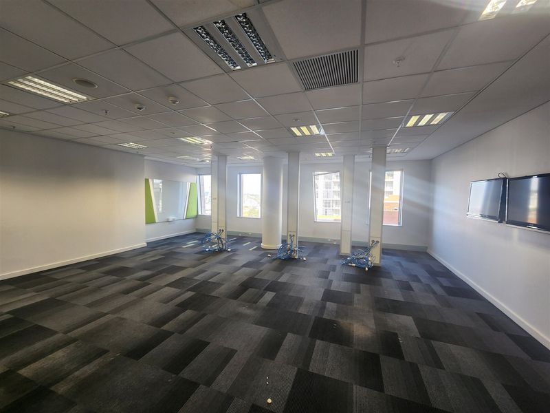 A spacious, vacant open-plan office featuring carpet tiles, suspended ceilings with recessed lighting, structural columns, and wall-mounted display screens.