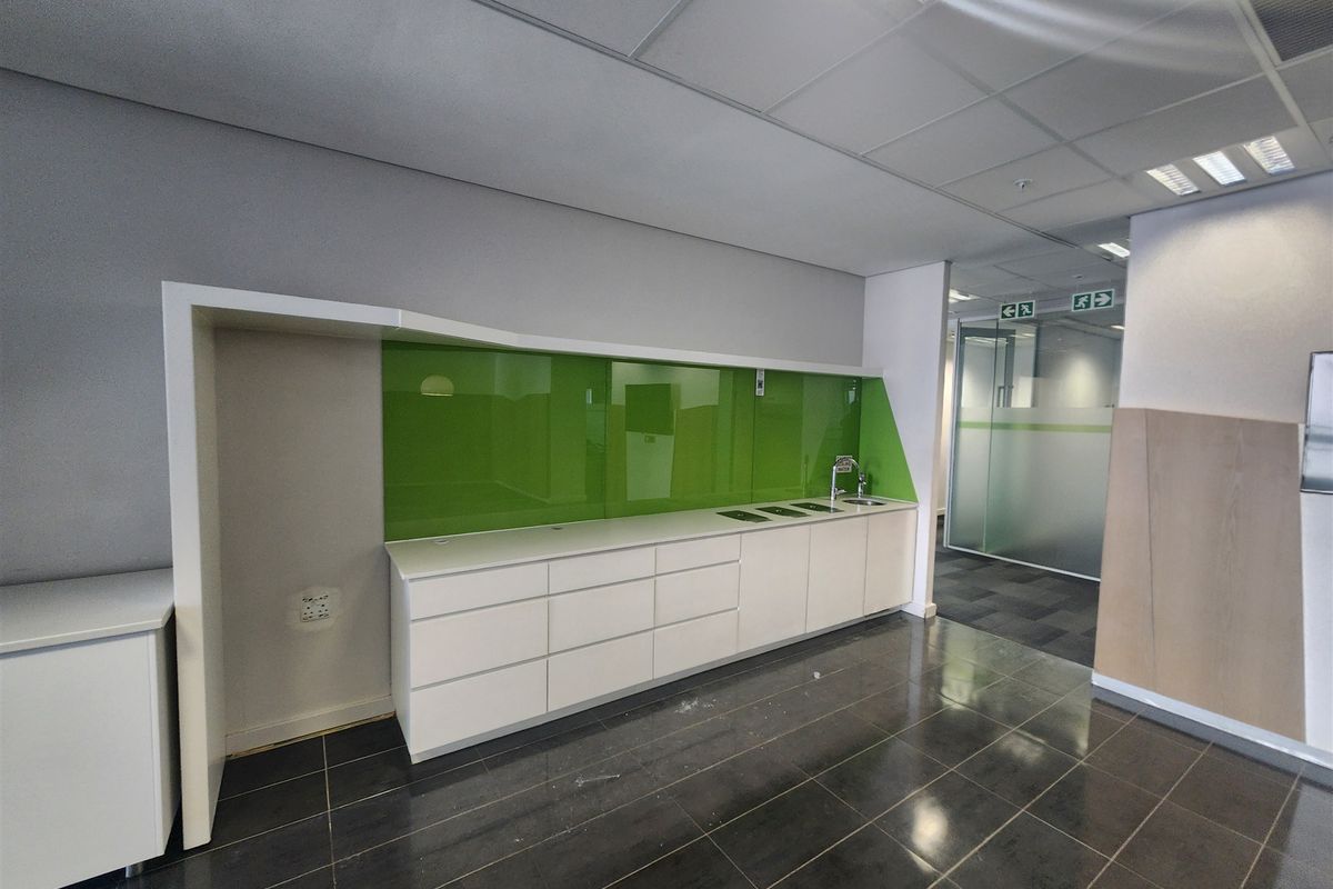 Modern office kitchenette featuring sleek white cabinetry, a vibrant green glass splashback, stainless steel sink, and durable dark tiled flooring.