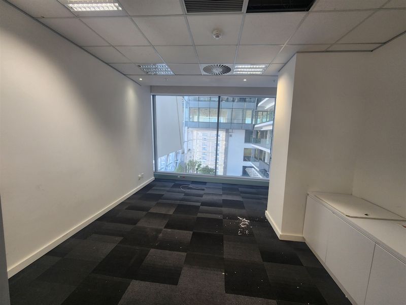 An empty office space featuring carpet tiles, built-in cabinetry, and a large window with views of the surrounding building complex.