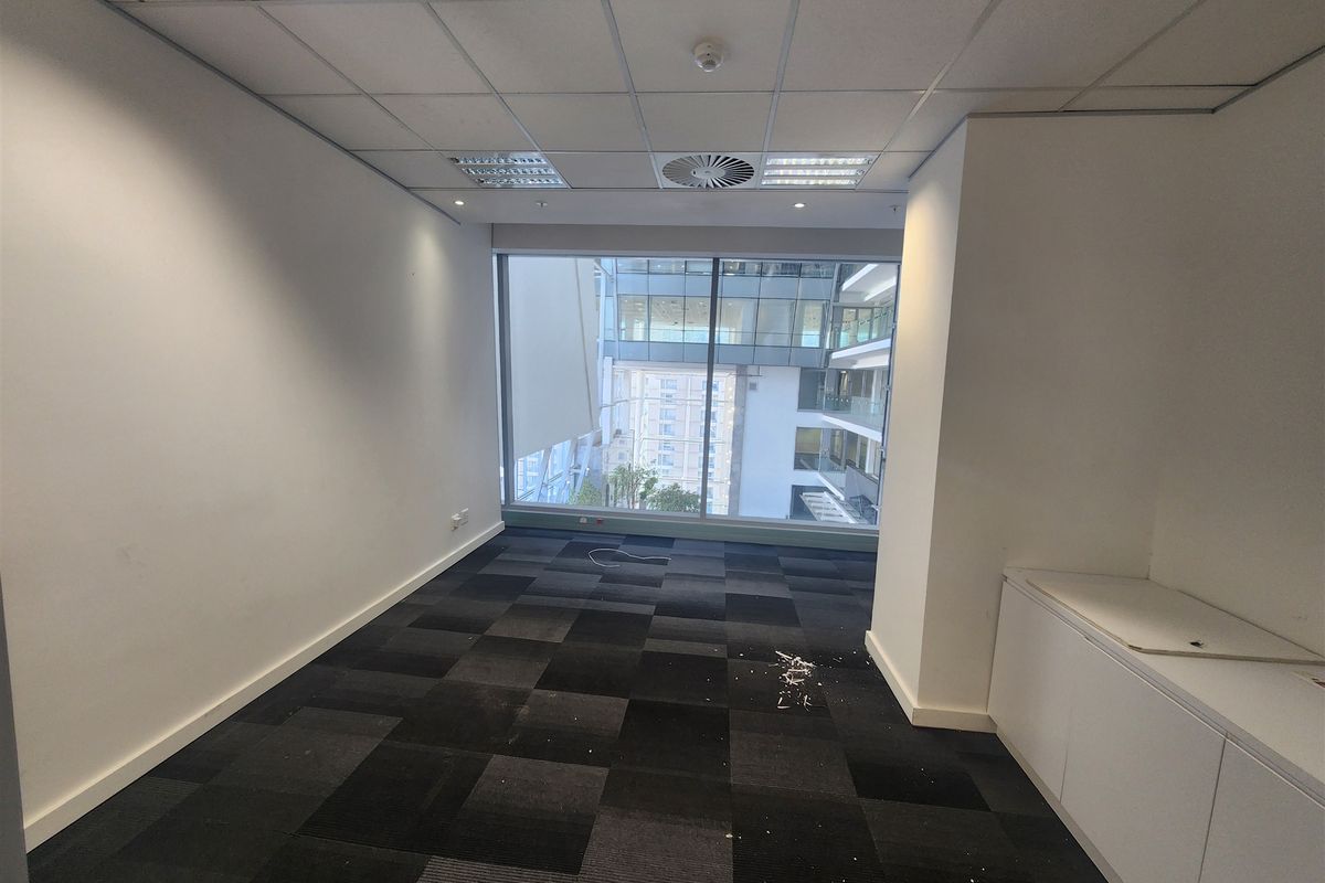An empty office space featuring carpet tiles, built-in cabinetry, and a large window with views of the surrounding building complex.