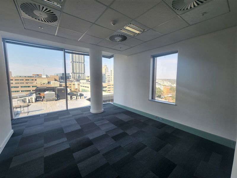 A bright, empty office space featuring floor-to-ceiling windows with city views, carpet tiles, and a modern suspended ceiling with integrated lighting and air conditioning.