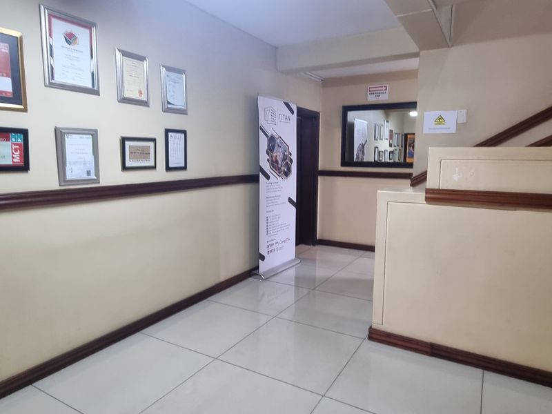 A clean, well-maintained office hallway featuring tiled flooring, a wooden dado rail, and a staircase balustrade.