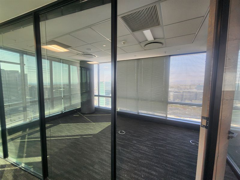 A modern, empty office space featuring floor-to-ceiling glass partitions, suspended ceilings with integrated lighting, and large windows with venetian blinds.