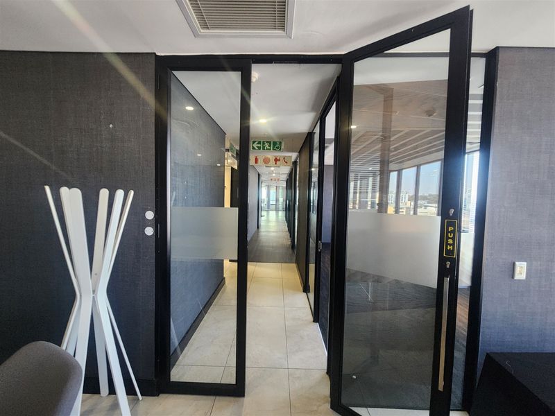 A modern office hallway featuring black-framed glass doors, tiled flooring, and recessed ceiling lighting with clear emergency exit signage.