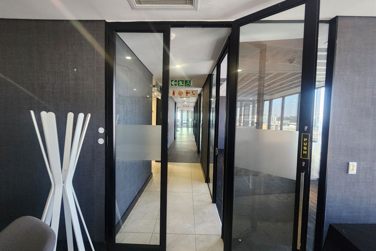 A modern office hallway featuring black-framed glass doors, tiled flooring, and recessed ceiling lighting with clear emergency exit signage.