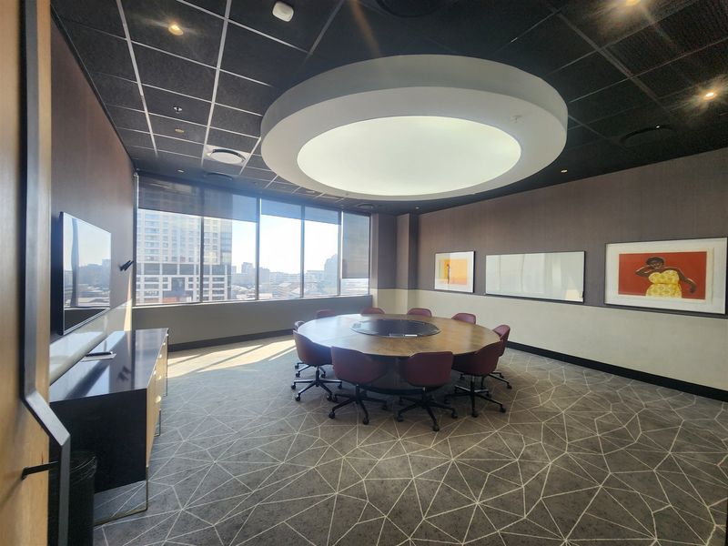 A modern, professional boardroom featuring a large circular feature light, floor-to-ceiling windows with city views, and high-quality finishes throughout.