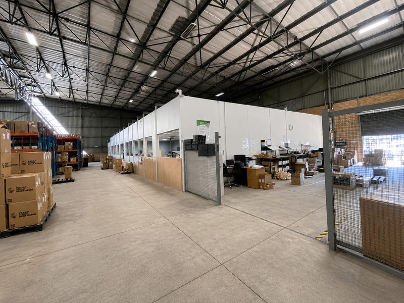 Spacious industrial warehouse featuring high exposed steel roof trusses, concrete flooring, and a partitioned office area with integrated security fencing.