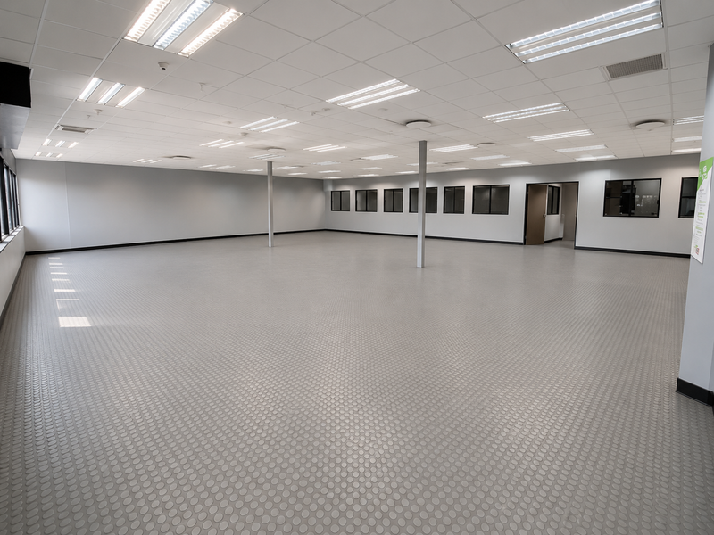 A spacious, open-plan industrial office or showroom area featuring durable rubber flooring, suspended ceilings with integrated lighting, and large windows.
