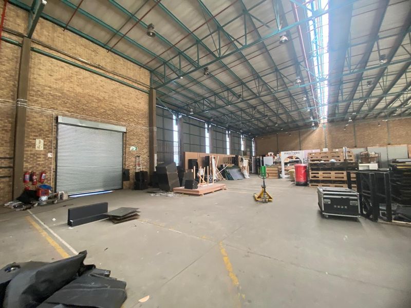 Spacious industrial warehouse unit featuring high exposed steel roof trusses, concrete flooring, brick walls, and a large roller shutter door for easy access.