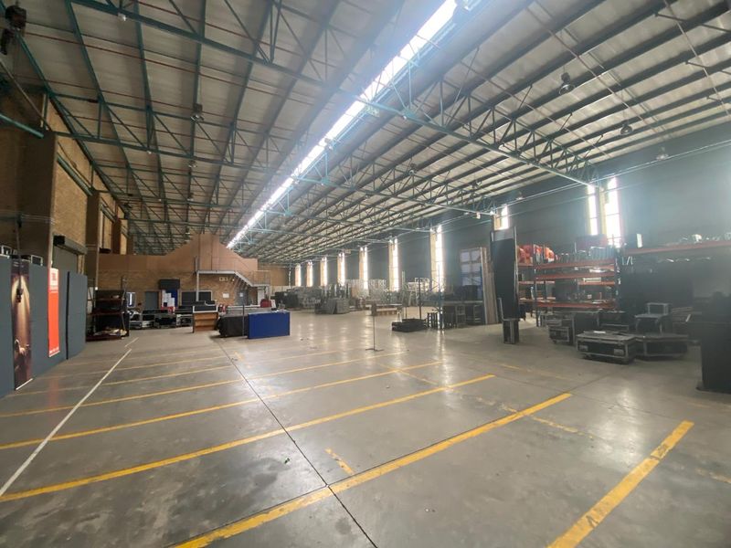 Spacious industrial warehouse featuring high ceilings with exposed steel trusses, concrete flooring, and natural light provided by overhead skylights.