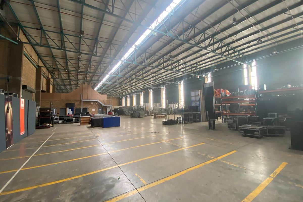 Spacious industrial warehouse featuring high ceilings with exposed steel trusses, concrete flooring, and natural light provided by overhead skylights.