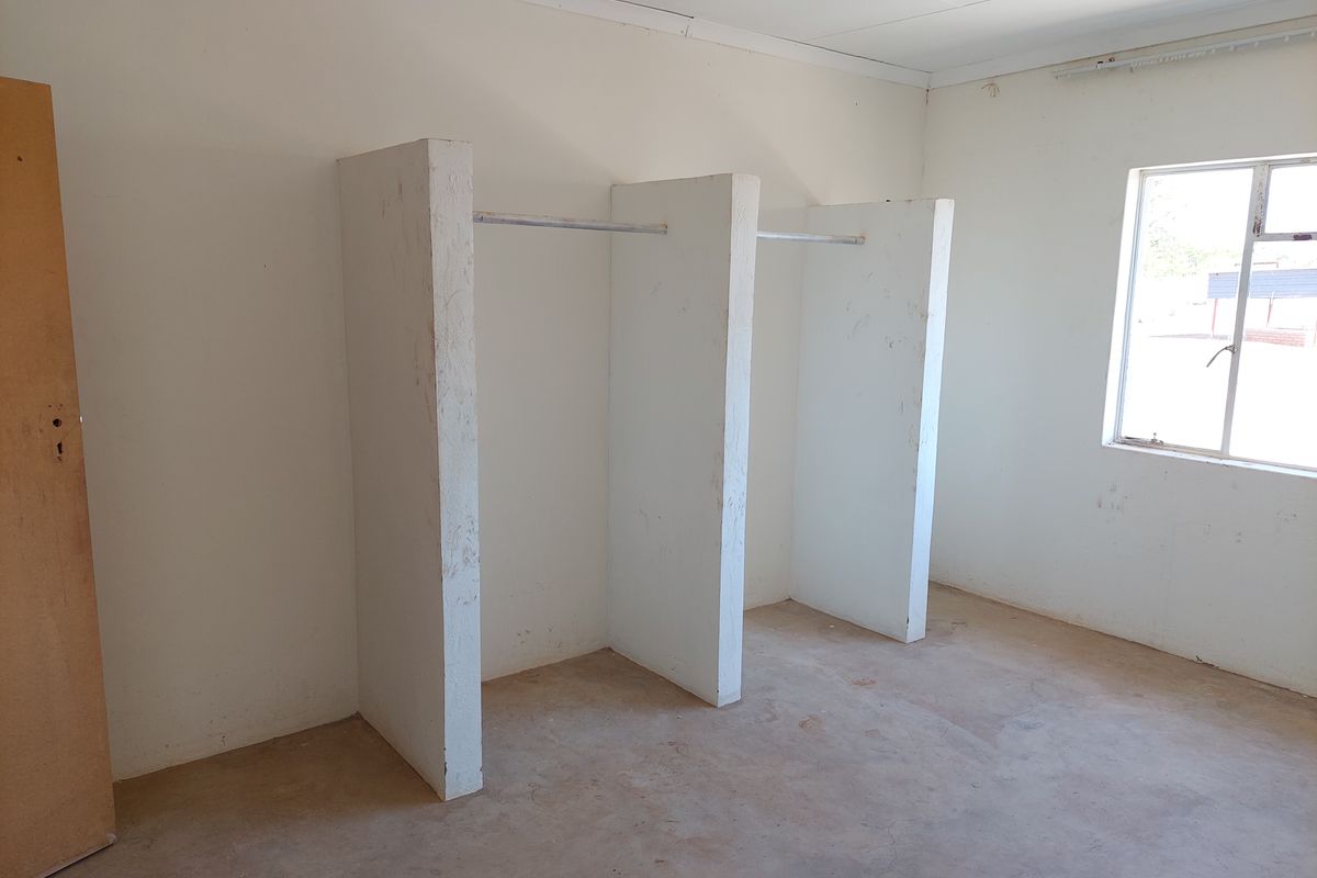 An empty bedroom featuring built-in wardrobe partitions with hanging rails and a window, requiring cosmetic updates to the walls and flooring.