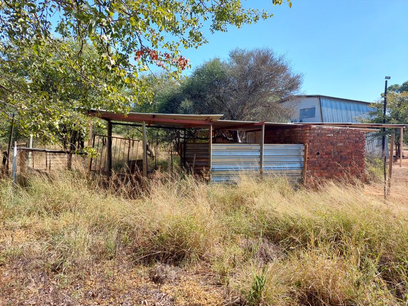 A rustic outdoor utility structure featuring brick masonry and corrugated iron roofing, situated in a natural farm setting with overgrown surroundings.