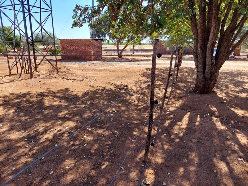 An outdoor farm area featuring an electric fence, a metal lattice tower, and a small brick outbuilding on dry, open ground.