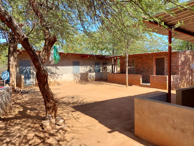 A rustic farm courtyard featuring exposed brick structures, covered patio areas, and natural sandy ground under mature trees.