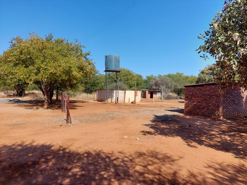A rural farm yard area featuring a water tank on a steel stand, a brick outbuilding, and open dirt grounds.
