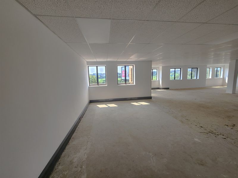 A large, open-plan office space featuring a suspended ceiling, recessed lighting, and multiple windows providing natural light, currently awaiting final flooring installation.