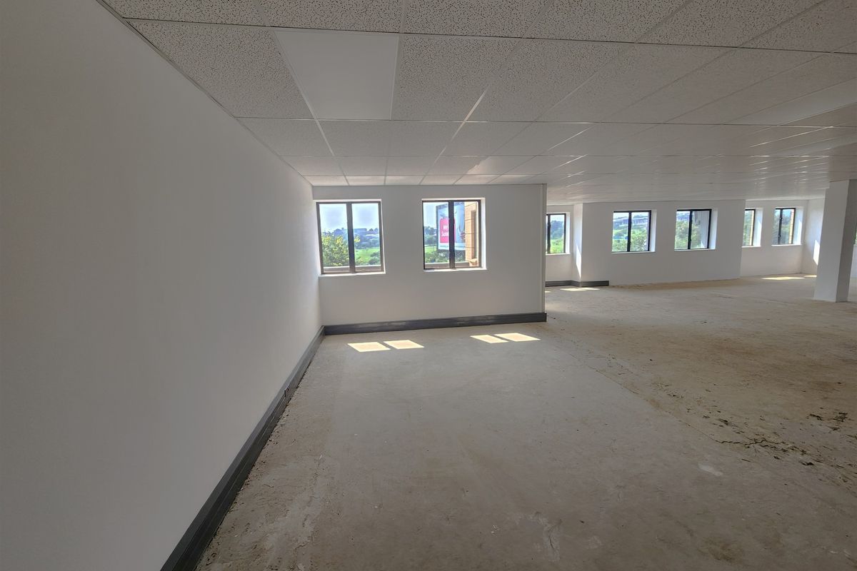A large, open-plan office space featuring a suspended ceiling, recessed lighting, and multiple windows providing natural light, currently awaiting final flooring installation.
