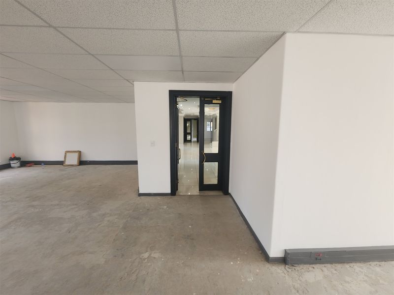 An empty office space featuring a suspended ceiling, unfinished concrete flooring, and a dark-framed glass door leading to an adjacent area.