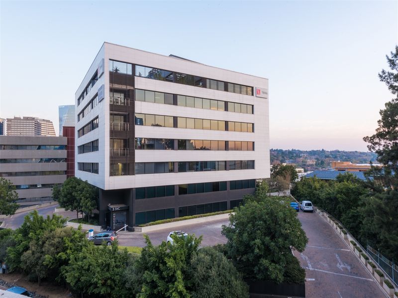 A professional aerial view of a modern multi-story office building featuring a clean facade, glass curtain walls, and a paved visitor entrance area.