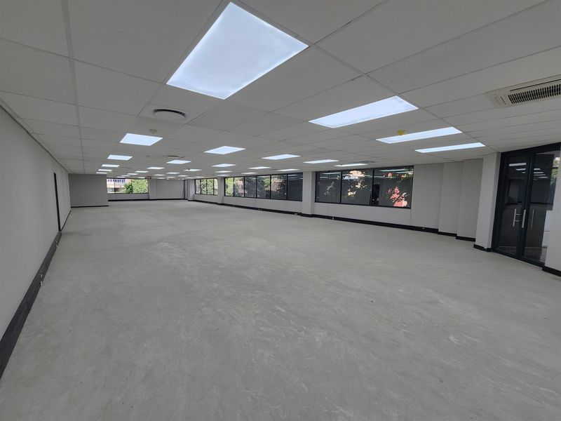 A spacious, open-plan office area featuring a suspended ceiling with recessed lighting, large windows for natural light, and integrated air conditioning.