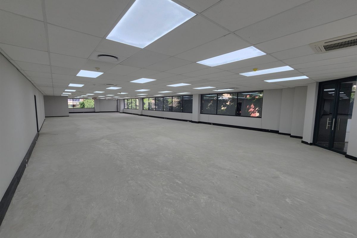 A spacious, open-plan office area featuring a suspended ceiling with recessed lighting, large windows for natural light, and integrated air conditioning.