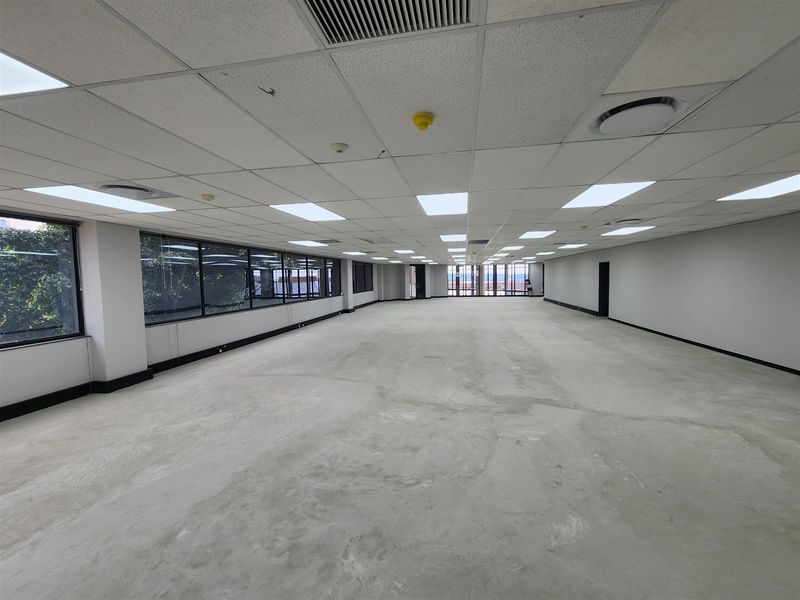 A large, open-plan office space featuring a suspended ceiling with recessed lighting, air conditioning vents, and expansive windows providing natural light.