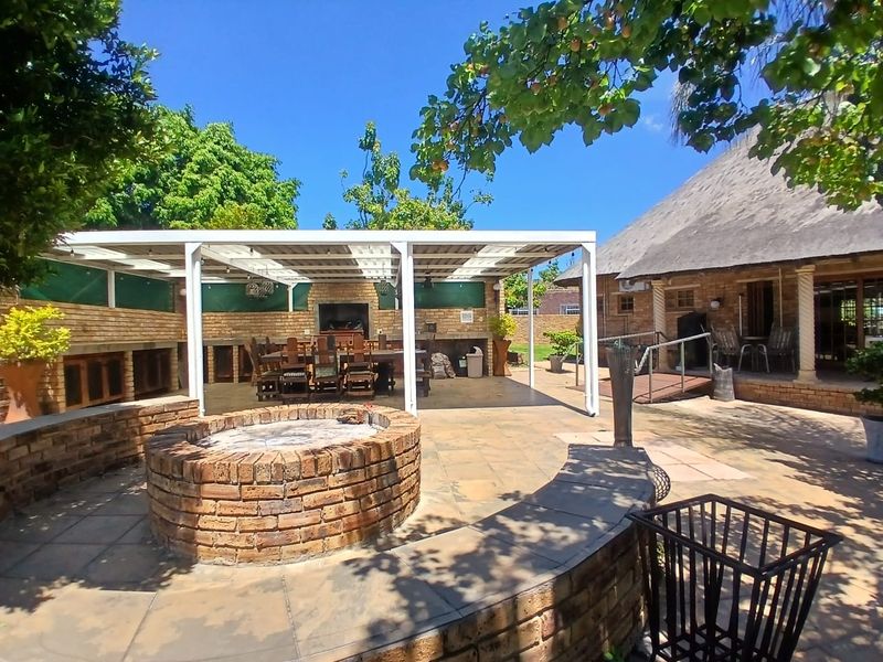Spacious outdoor entertainment area featuring a covered patio with a built-in braai and a circular brick fire pit in a landscaped garden setting.