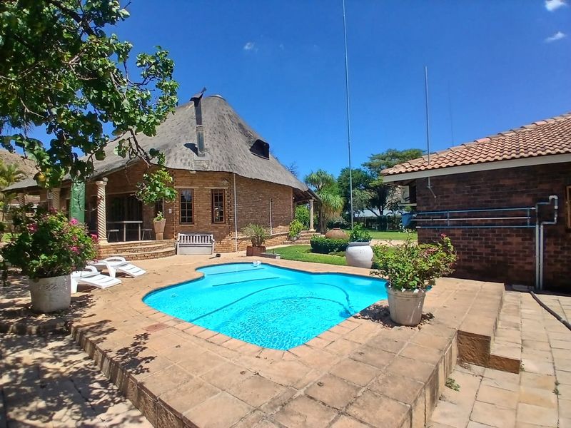Outdoor pool area featuring a swimming pool with tiled surround, set against a traditional thatched-roof dwelling and brick outbuilding.
