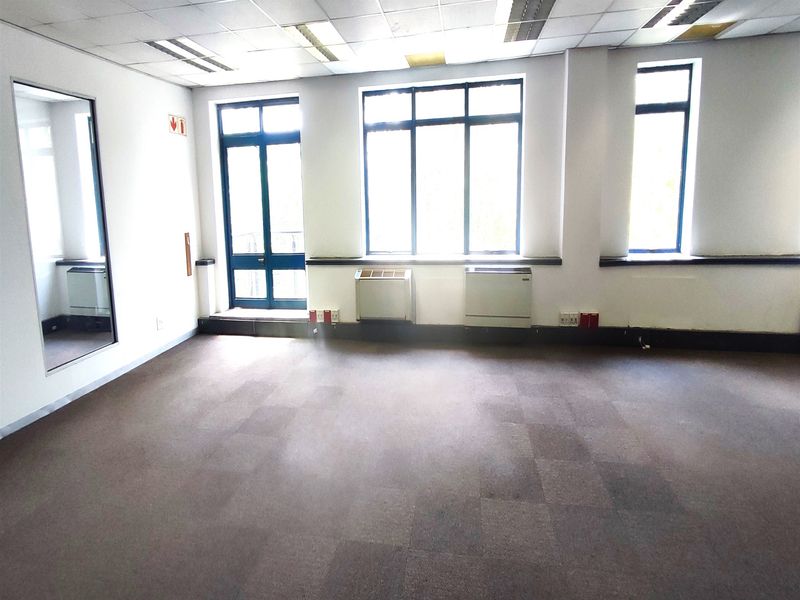 An empty, open-plan office space featuring carpeted flooring, suspended ceilings with recessed lighting, and large windows providing natural light.