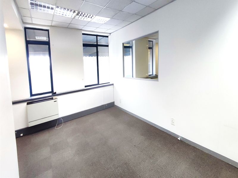 A clean, empty office space featuring carpet flooring, a suspended ceiling with recessed lighting, an internal window, and a wall-mounted HVAC unit.